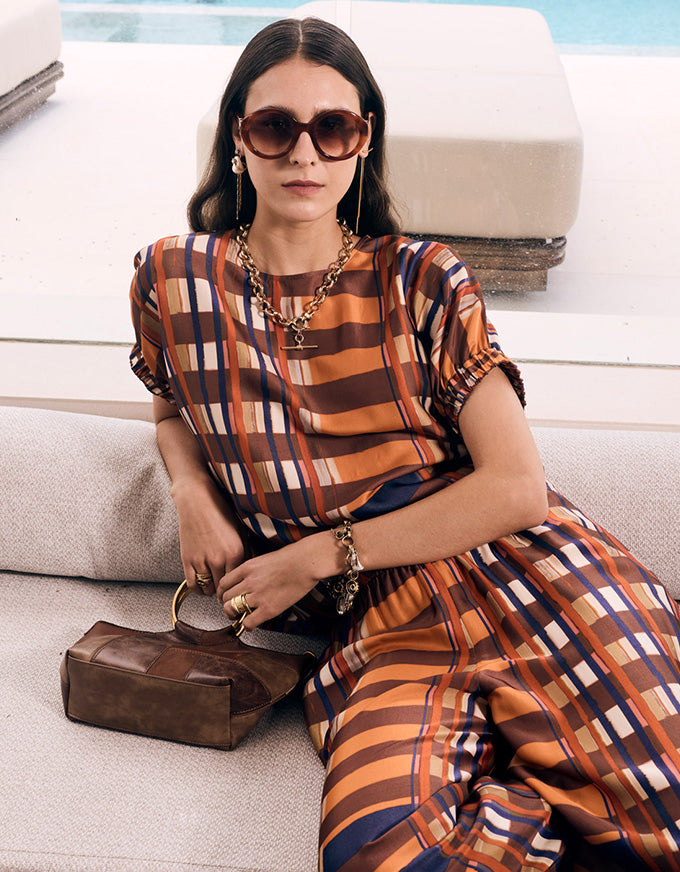 A woman wearing a brown, orange, and cream striped dress sits on a light-colored outdoor sofa. She is styled with brown sunglasses, a chunky gold chain necklace, a gold bracelet, and a brown leather handbag.