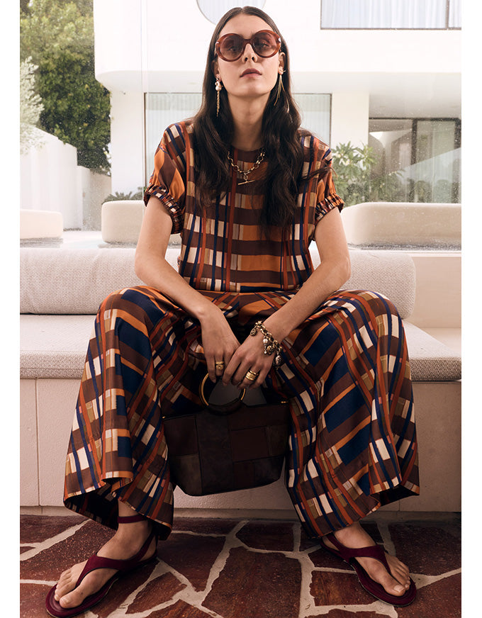 A woman sits on a modern outdoor bench wearing a matching brown, orange, and navy plaid short-sleeve top and wide-leg pants set. She is styled with sunglasses, a dark brown handbag, and burgundy flip-flops.