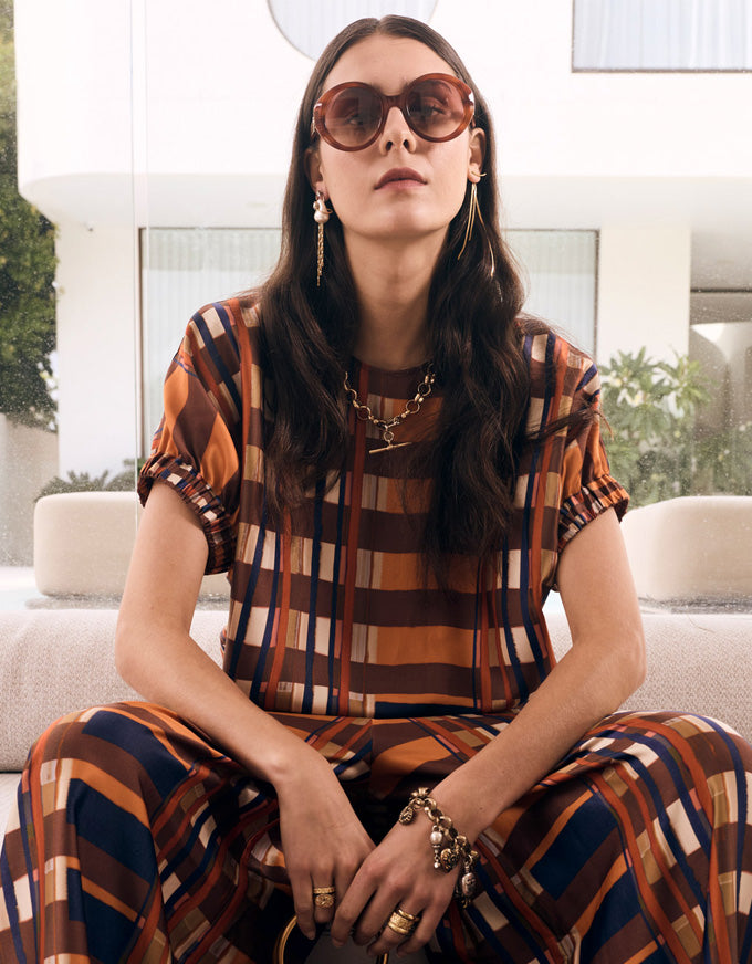 A woman wearing a matching brown, orange, and navy plaid short-sleeve top and trousers set, styled with sunglasses, gold jewelry, and seated on a light-colored sofa in a modern outdoor setting.
