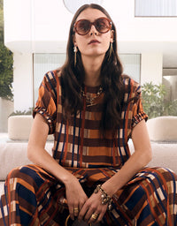 A woman wearing a matching brown, orange, and navy plaid short-sleeve top and trousers set, styled with sunglasses, gold jewelry, and seated on a light-colored sofa in a modern outdoor setting.