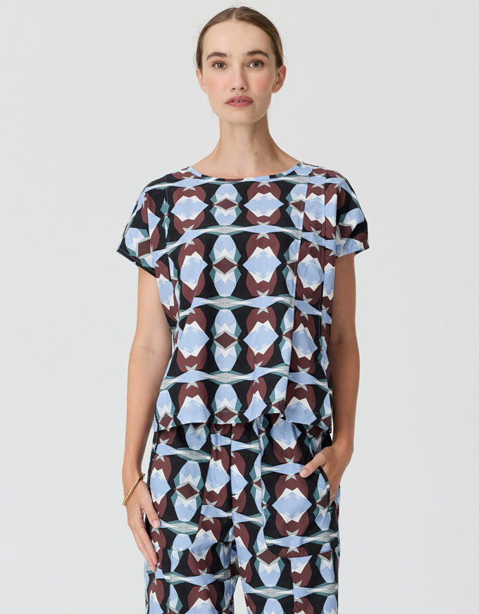 A woman modeling a short-sleeve jumpsuit with a geometric pattern in light blue, white, and dark brown on a light gray background.