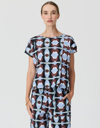 A woman modeling a short-sleeve jumpsuit with a geometric pattern in light blue, white, and dark brown on a light gray background.