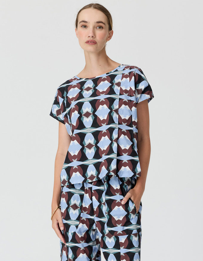 A woman modeling a matching short-sleeve top and trousers set with a geometric abstract print in light blue, brown, and black on a light gray background.