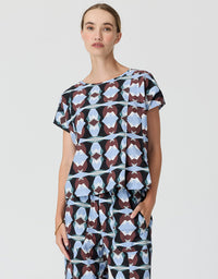 A woman modeling a matching short-sleeve top and trousers set with a geometric abstract print in light blue, brown, and black on a light gray background.