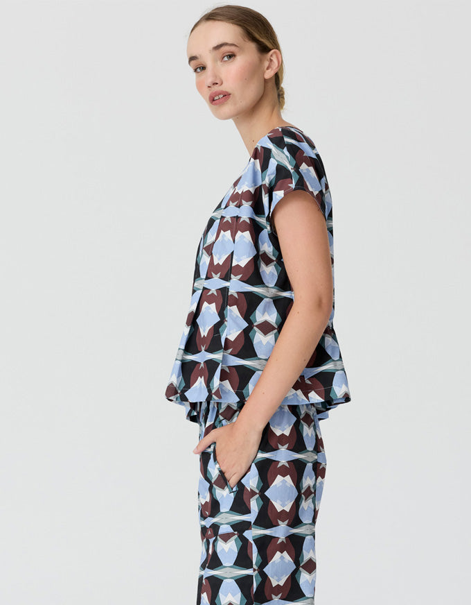 A woman modeling a matching sleeveless top and trousers set with a geometric abstract print in light blue, white, and dark brown on a light gray background.