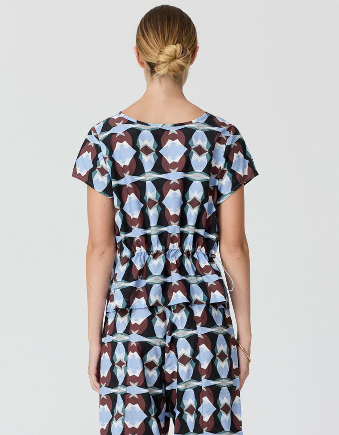 A woman viewed from the back wearing a short-sleeve jumpsuit with a geometric pattern in light blue, brown, and black on a light gray studio background.