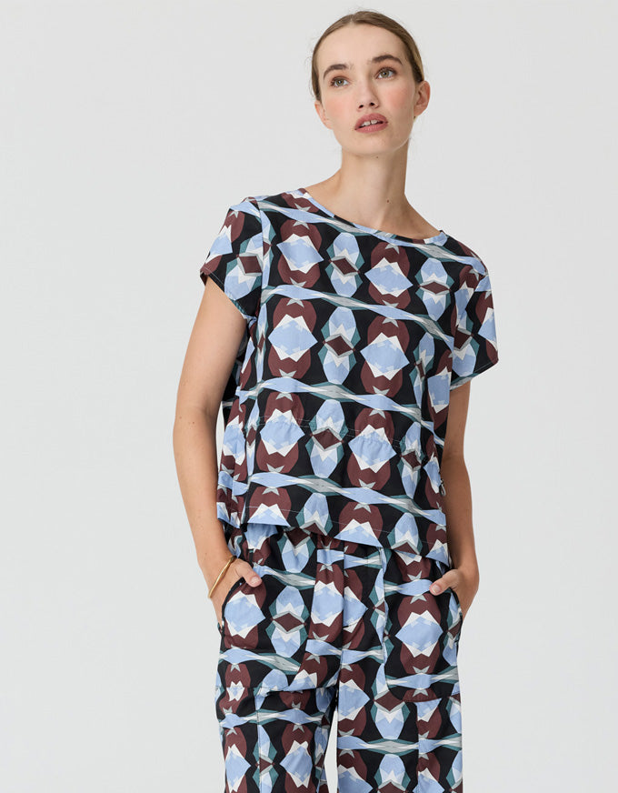 A woman modeling a matching short-sleeve top and trousers set with a geometric abstract pattern in light blue, dark brown, and black, set against a plain light gray background.