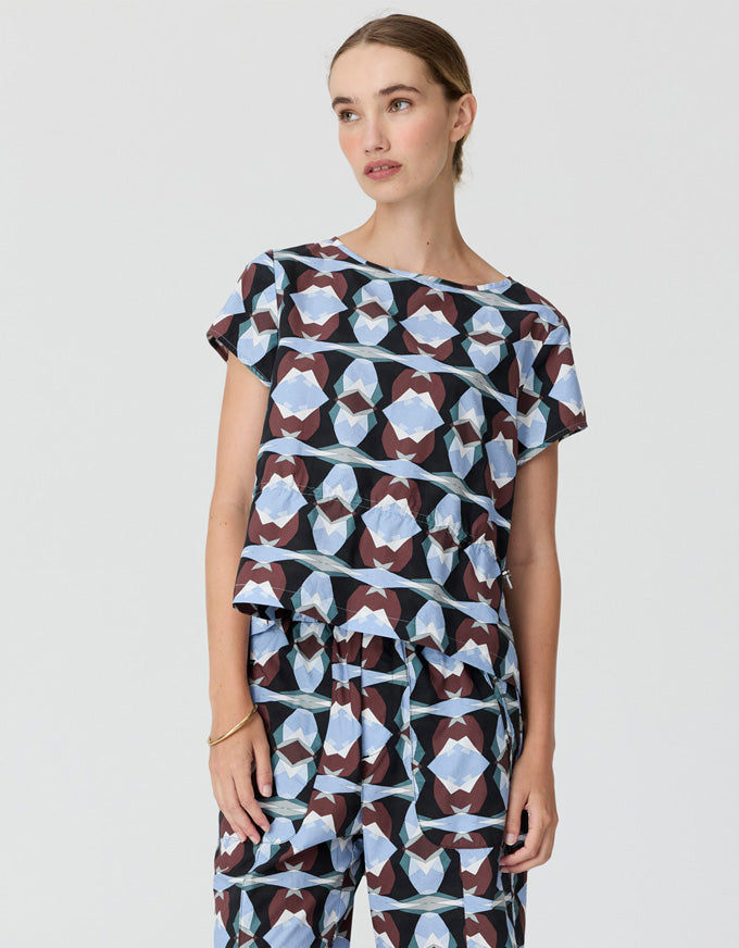 A woman modeling a matching short-sleeve top and wide-leg pants set with an abstract geometric pattern in light blue, white, and dark brown on a black background, photographed against a light gray studio backdrop.