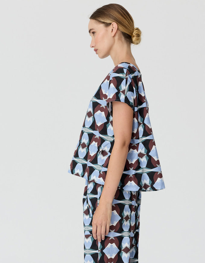A woman shown in side profile wearing a matching short-sleeve top and trousers set with a geometric abstract print in light blue, white, and dark brown on a light gray background.