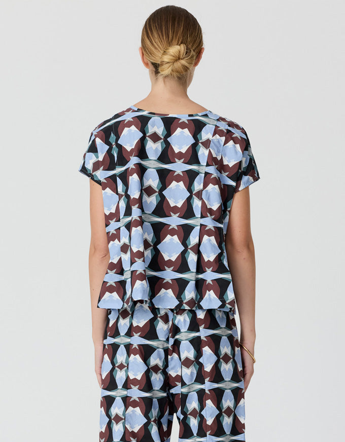 A woman viewed from the back wearing a matching short-sleeve top and pants set with an abstract geometric pattern in light blue, brown, and white on a dark background, set against a plain light gray studio backdrop.