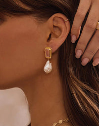 Close-up of a woman wearing a gold earring with a pearl drop, with her hand touching her hair.