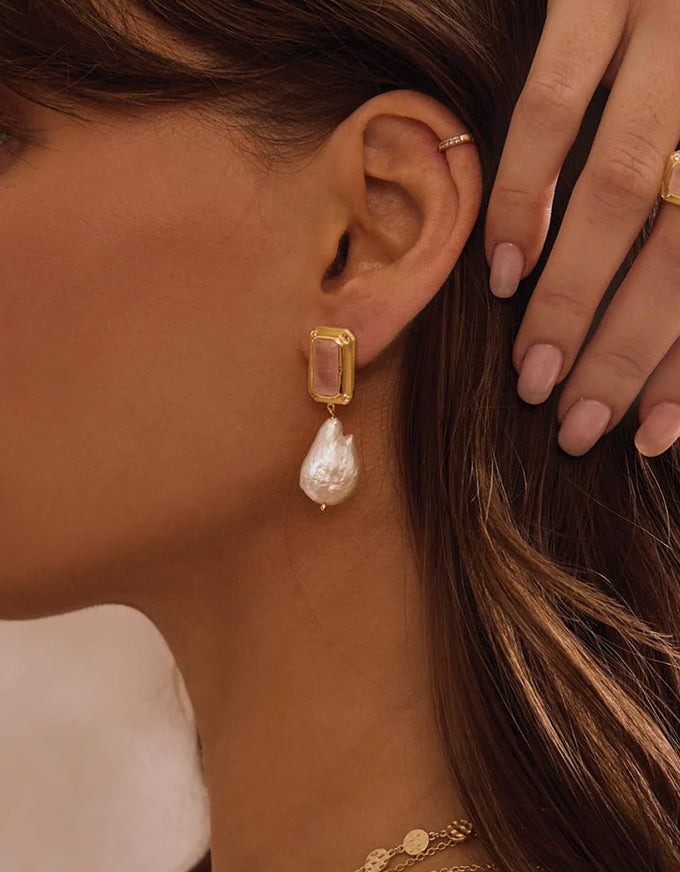 Close-up of a woman wearing a gold earring with a pearl drop, with her hand touching her hair.