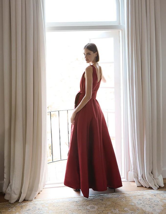 Woman in a red dress standing in a room with large windows and white curtains.