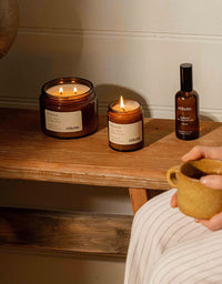 Two lit candles and a bottle of essential oil on a wooden surface with a person holding a mug.
