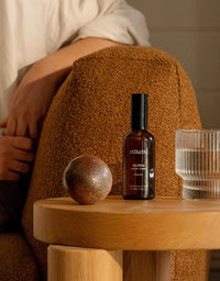 Bottle of etikette skincare product on a wooden stool with a textured brown cushion and glass in the background.