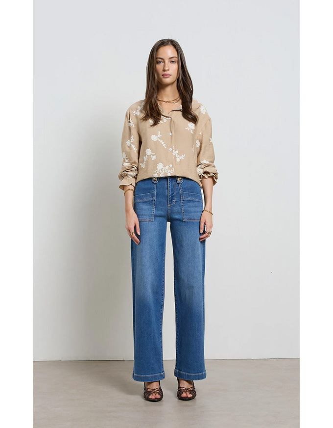 Woman wearing a beige floral blouse and blue jeans against a plain background