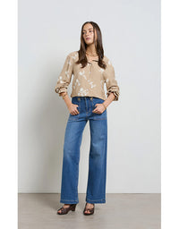 Woman wearing a beige floral blouse and blue jeans against a plain background