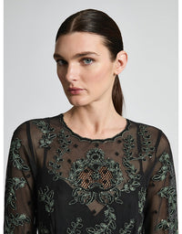 Woman wearing a black sheer top with intricate green embroidery on a white background