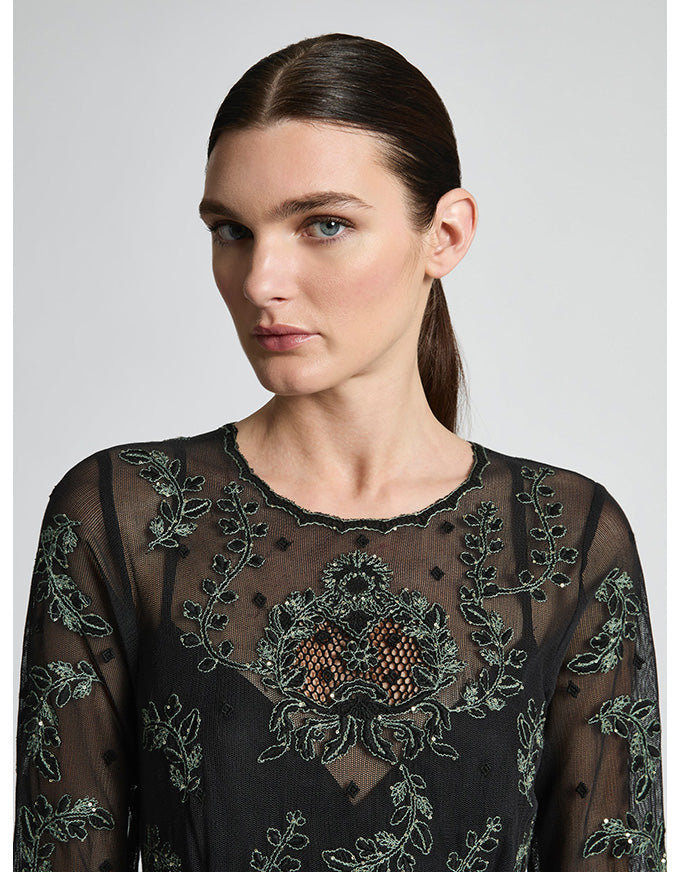 Woman wearing a black sheer top with intricate green embroidery on a white background