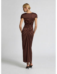 A woman modeling a floor-length brown satin dress from the back, featuring short cap sleeves, a boat neckline, and a gathered ruched back seam, paired with black strappy heels against a light gray studio background.