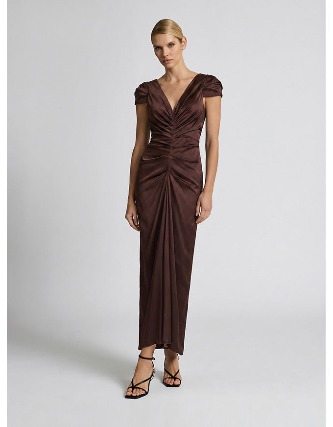 A woman modeling a floor-length, dark brown ruched gown with cap sleeves and a deep V-neckline, paired with black strappy heels, against a light gray studio background.
