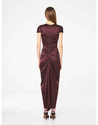 A woman viewed from the back wearing a floor-length draped satin dress in a deep plum-burgundy color, paired with black high heels, against a plain white studio background.