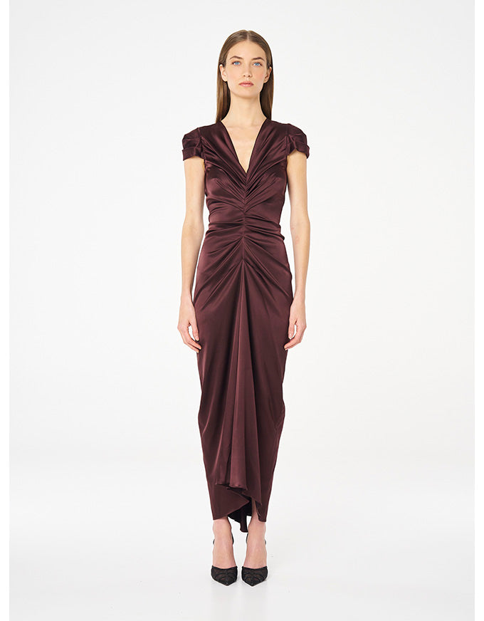 A woman modeling a floor-length, deep V-neck dress in a rich burgundy satin. The dress features cap sleeves, a gathered ruched bodice, and a draped skirt, paired with black pointed-toe heels against a plain white studio background.