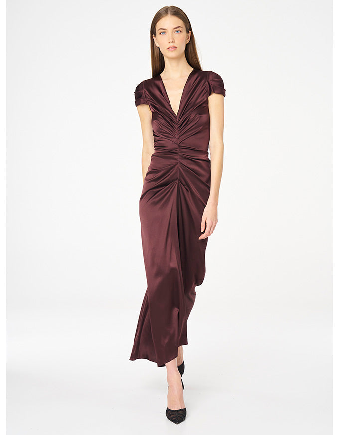 A woman modeling a floor-length, deep V-neck satin dress in a dark burgundy color, featuring cap sleeves and a gathered ruched bodice, set against a plain white studio background.