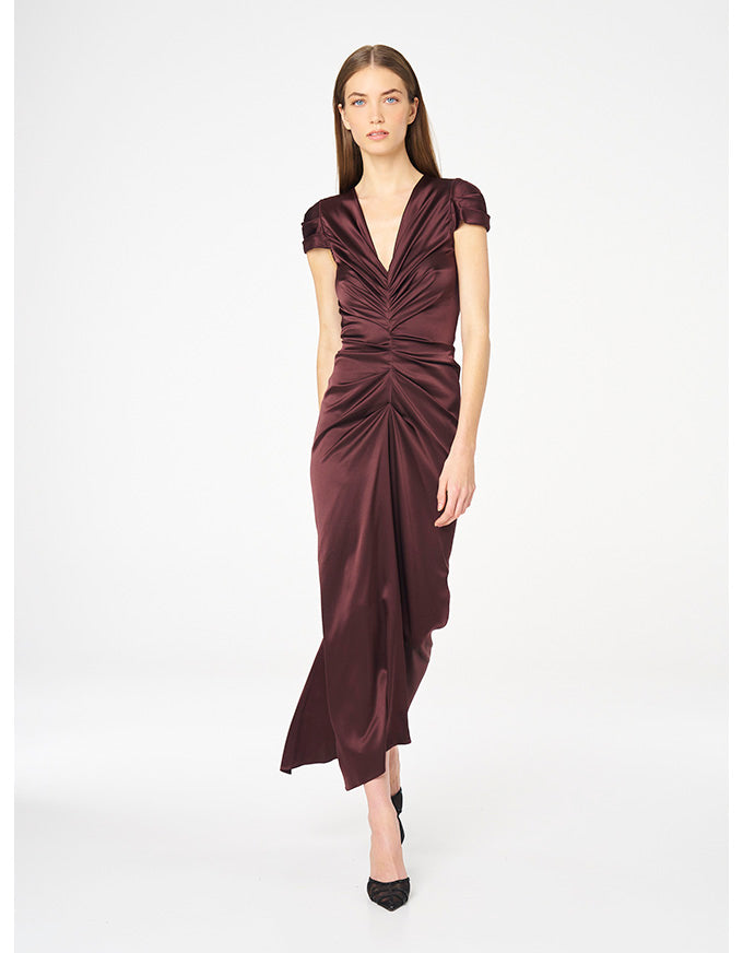 A woman modeling a floor-length draped satin dress in deep burgundy with short cap sleeves and a gathered waist, photographed against a clean white studio background.