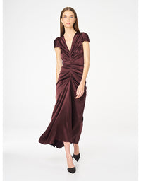A woman models a floor-length, deep V-neck dress in a rich burgundy satin. The dress features short cap sleeves, a gathered ruched bodice, and a flowing skirt, styled with black pointed-toe heels against a plain white studio background.