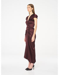 A woman modeling a dark plum satin midi dress with a draped, gathered waist and short cap sleeves, paired with black pointed-toe heels, against a plain white studio background.
