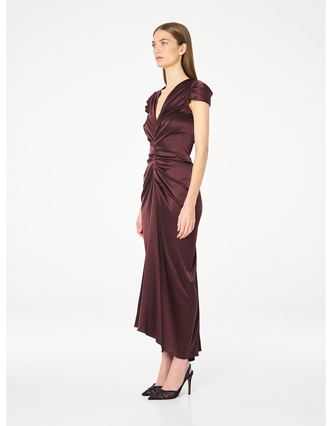 A woman modeling a dark plum satin midi dress with a draped, gathered waist and short cap sleeves, paired with black pointed-toe heels, against a plain white studio background.