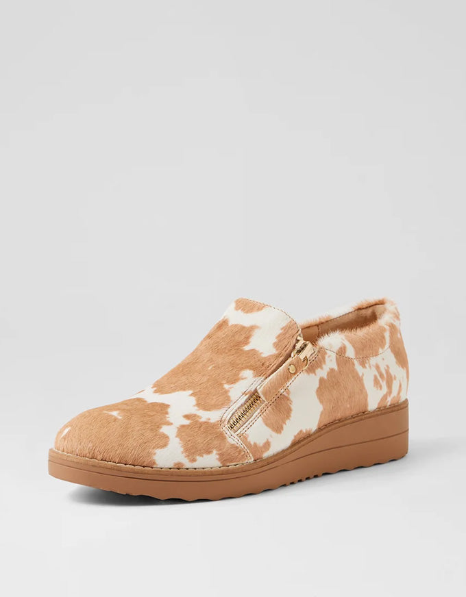 Tan and white cow print slip-on shoe with a brown sole on a light gray background