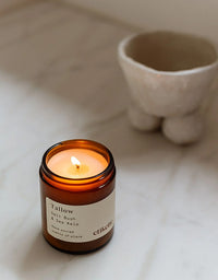 Candle in an amber jar labeled 'Tallow' on a light surface with a blurred ceramic pot in the background.