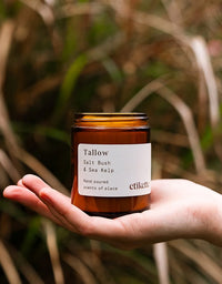 Hand holding a 'Tallow' candle labeled 'Salt Bush & Sea Kelp' by 'etikette' against a natural background.