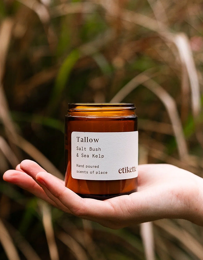 Hand holding a 'Tallow' candle labeled 'Salt Bush & Sea Kelp' by 'etikette' against a natural background.