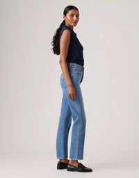 A woman stands in profile wearing light-wash blue jeans, a dark navy sleeveless top, and black loafers against a plain light gray studio background.