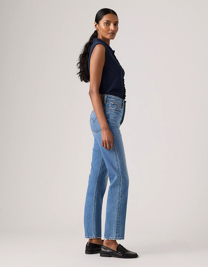 A woman stands in profile wearing light-wash blue jeans, a dark navy sleeveless top, and black loafers against a plain light gray studio background.