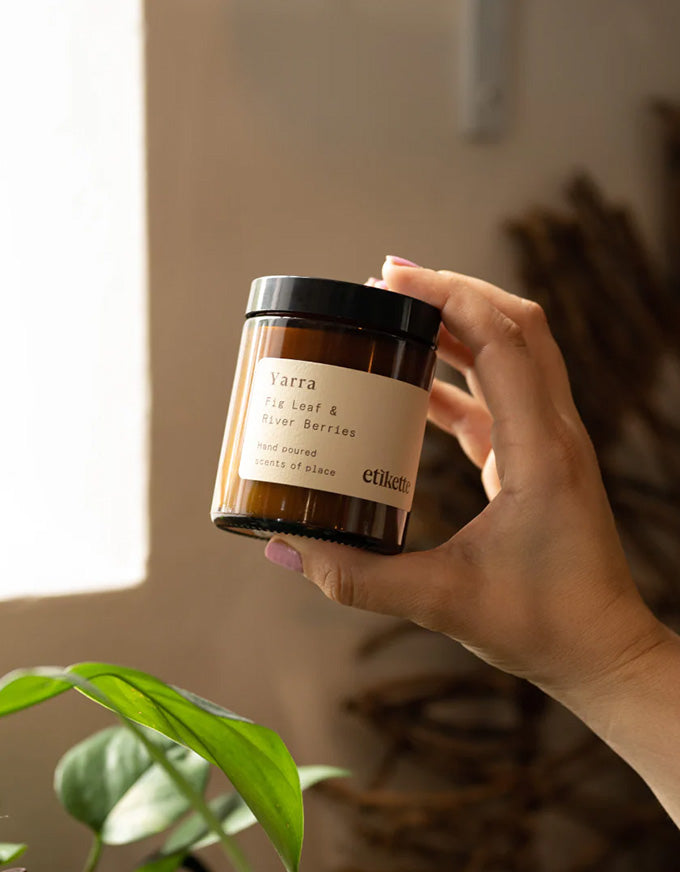 Hand holding a candle jar labeled 'Verra' with a blurred indoor background