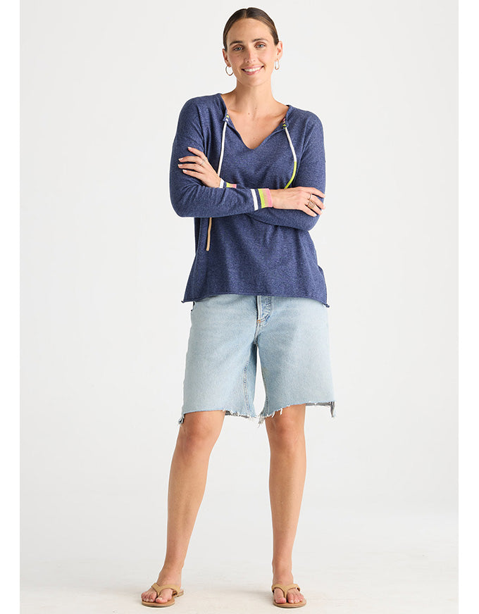 A woman modeling a navy blue long-sleeve V-neck top with light blue denim shorts and tan sandals, standing against a plain light gray studio background.