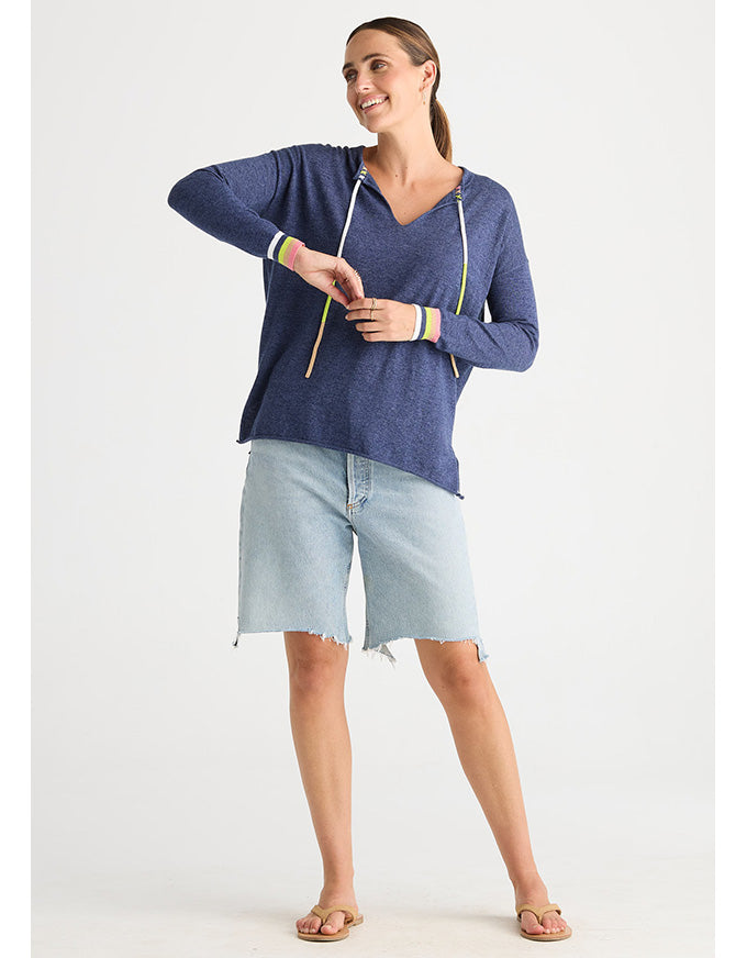 A woman modeling a heathered blue long-sleeve hoodie with colorful striped cuffs and light-wash denim shorts against a plain white studio background.