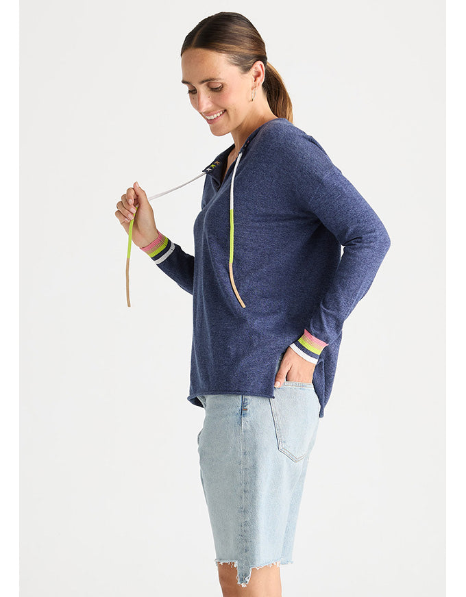 A woman wearing a navy blue heathered hoodie with green and white striped cuffs, paired with a light wash denim skirt, photographed against a solid off-white background.