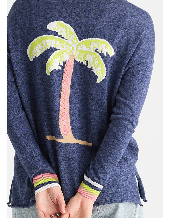 A person wearing a navy blue knit sweater with a large palm tree motif on the back in light green, pink, beige, and white, with striped ribbed cuffs in pink, green, white, and navy.
