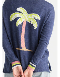 A person wearing a navy blue knit sweater with a large palm tree motif on the back in light green, pink, beige, and white, with striped ribbed cuffs in pink, green, white, and navy.