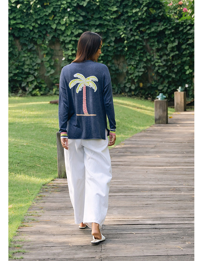 A woman viewed from behind walking on a wooden boardwalk wearing a navy blue long-sleeve top with a large embroidered palm tree on the back, paired with white wide-leg pants.