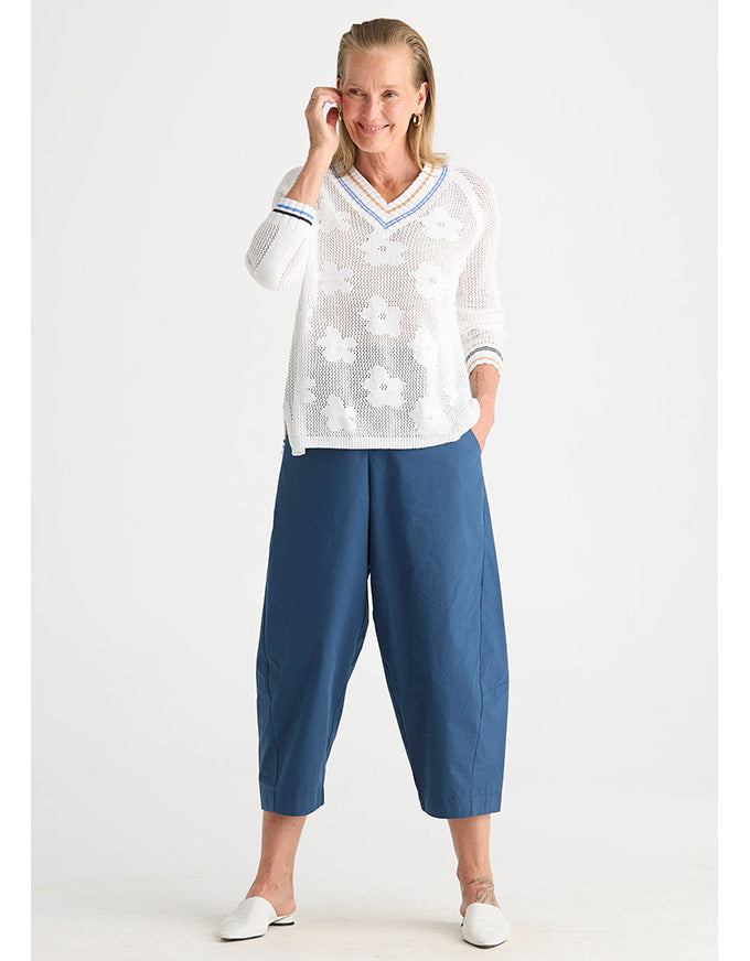 A woman modeling a white long-sleeve knit sweater with a V-neck, white floral embroidery, and blue striped trim, paired with blue cropped pants and white shoes, against a light gray studio background.