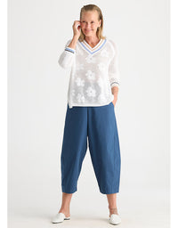 A woman modeling a white long-sleeve knit sweater with a V-neck, white floral embroidery, and blue striped trim, paired with blue cropped pants and white shoes, against a light gray studio background.