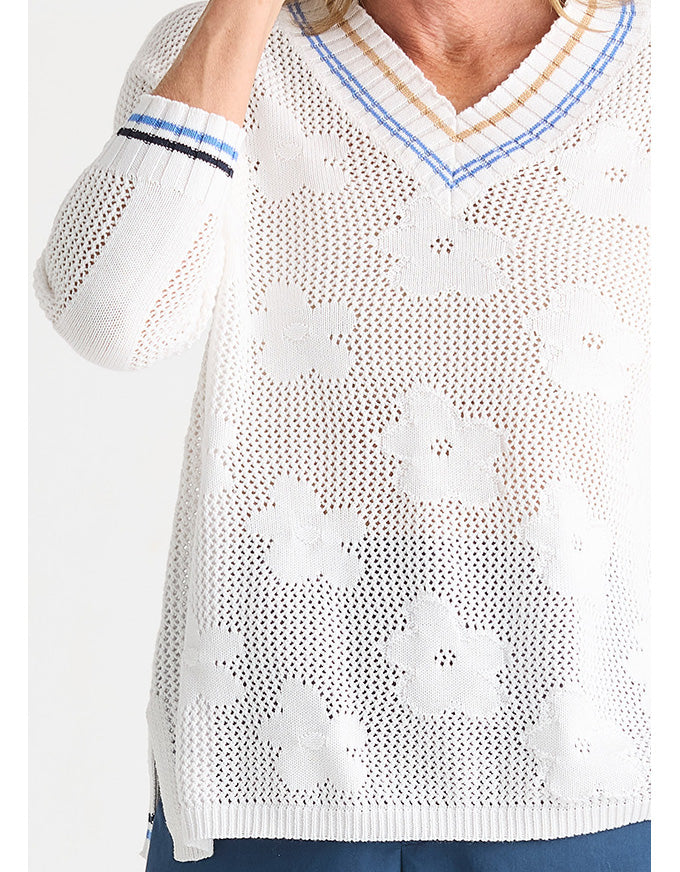 A close-up of a white knit sweater with a V-neck, featuring large floral lace patterns and striped trim in blue, beige, and white on the collar and cuffs.