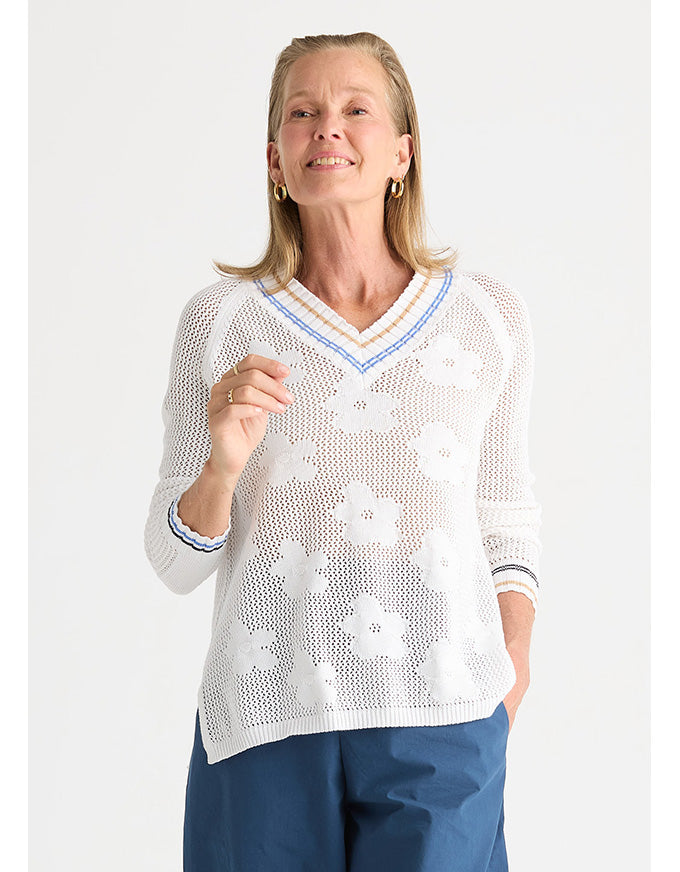 A woman wearing a white open-knit V-neck sweater with blue and white striped trim, paired with blue pants, against a light gray studio background.