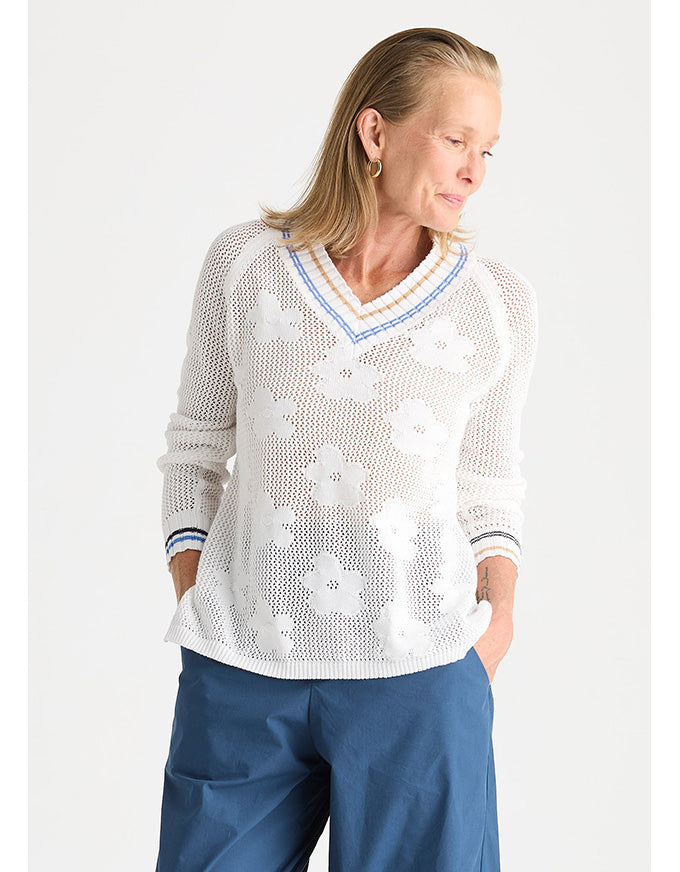 A woman wearing a white open-knit long-sleeve sweater with a blue and white V-neck trim, paired with blue pants, against a light gray studio background.